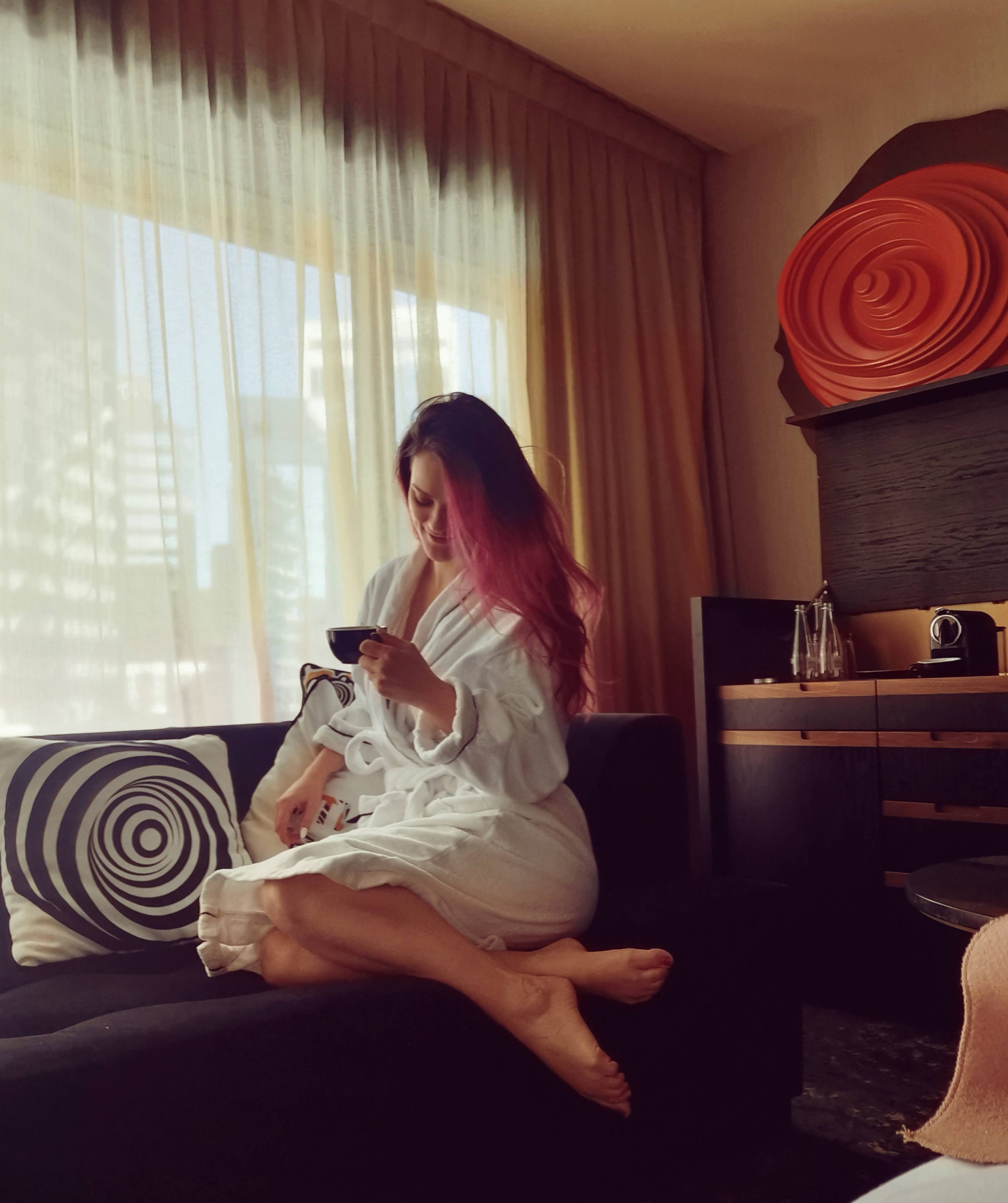 CyberCandace sitting by a window in a hotel room wearing a white robe, holding a black teacup with soft natural light.
